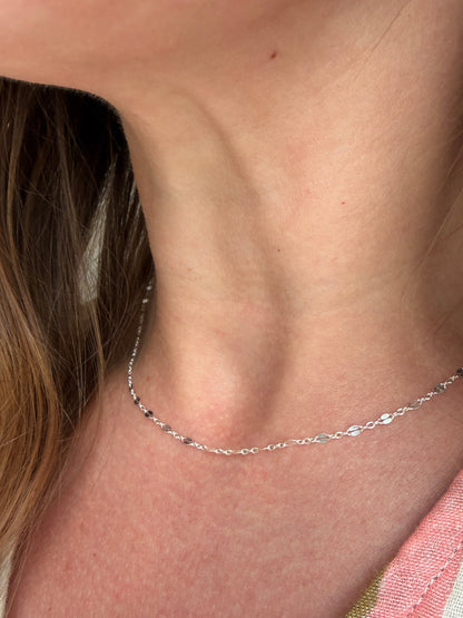 A close-up of a delicate silver chain necklace worn around a person's neck, with light brown hair and a pink and white striped shirt visible.