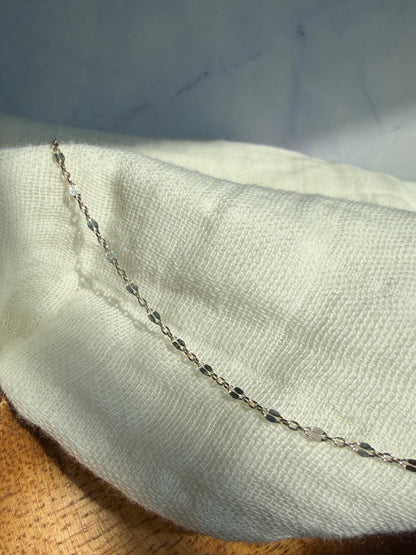 A close-up of a delicate silver chain draped over a textured off-white fabric, with a wooden surface visible in the background.