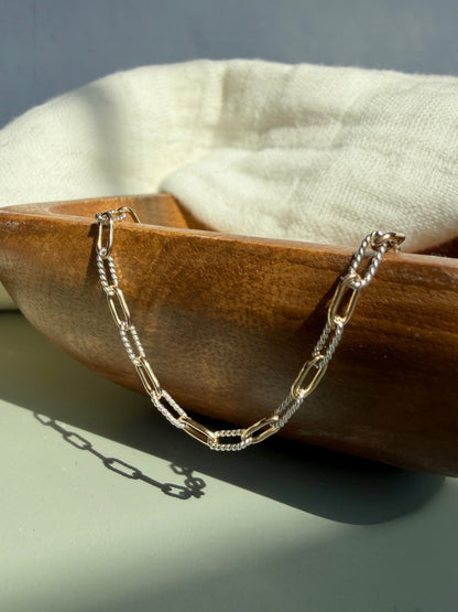 A silver paperclip chain necklace draped over a wooden bowl with a white fabric lining, photographed in warm natural light.