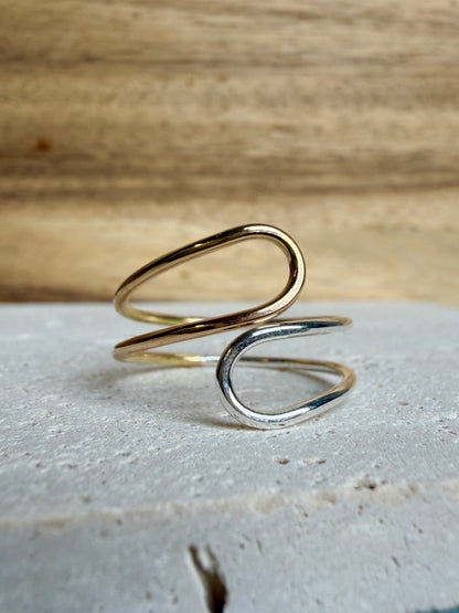 A minimalist two-tone spiral ring with one gold-toned loop and one silver-toned loop, displayed on a textured white stone slab with a wooden background.
